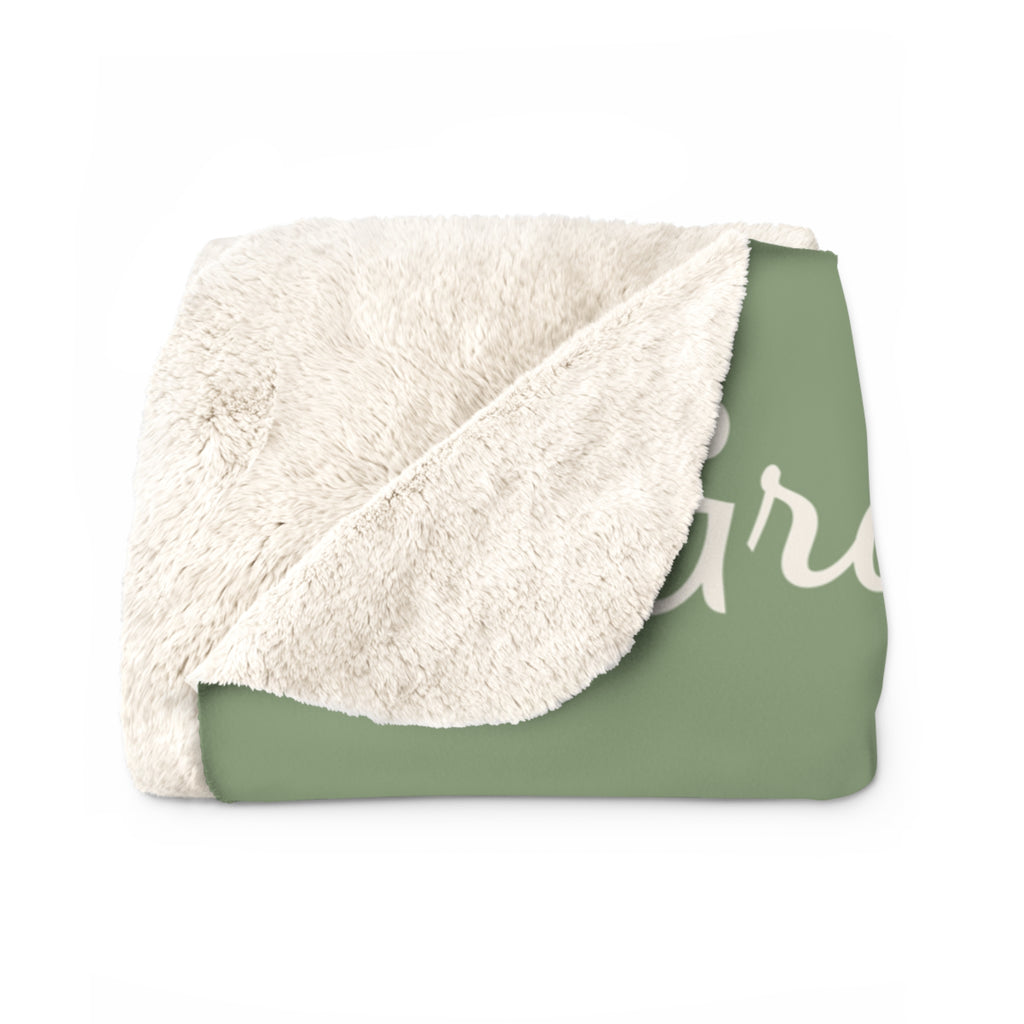 A folded Sage blanket for grandmother in sherpa fleece has one corner turned back to show its fluffy cream back beside the bouquet.