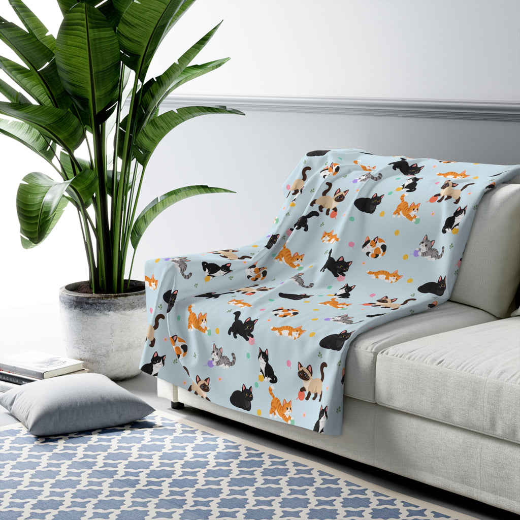 A Sky cat sherpa blanket drapes over a light gray sofa, with cartoon cats, pastel yarn balls and daisies on the fleece front.