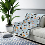 A Sky cat sherpa blanket drapes over a light gray sofa, with cartoon cats, pastel yarn balls and daisies on the fleece front.