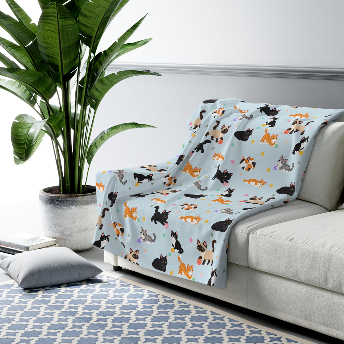 A Sky cat sherpa blanket drapes over a light gray sofa, with cartoon cats, pastel yarn balls and daisies on the fleece front.