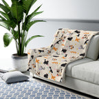 Cartoon cats cover a Cream sherpa fleece blanket draped over a light gray sofa in a bright room with a potted plant and patterned rug.