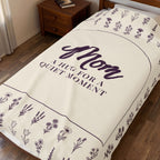 Cream velveteen plush hug blanket spread over a dark wooden single bed, showing "Mom" in a deep plum arch surrounded by wildflower rows.