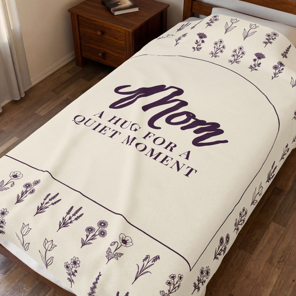 Cream velveteen plush hug blanket spread over a dark wooden single bed, showing "Mom" in a deep plum arch surrounded by wildflower rows.