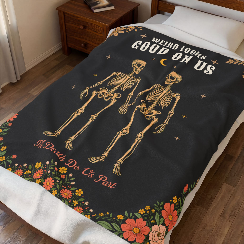 Seen from above, a Black velveteen plush blanket shows skeleton lovers holding hands on a made bed beside a wooden nightstand.