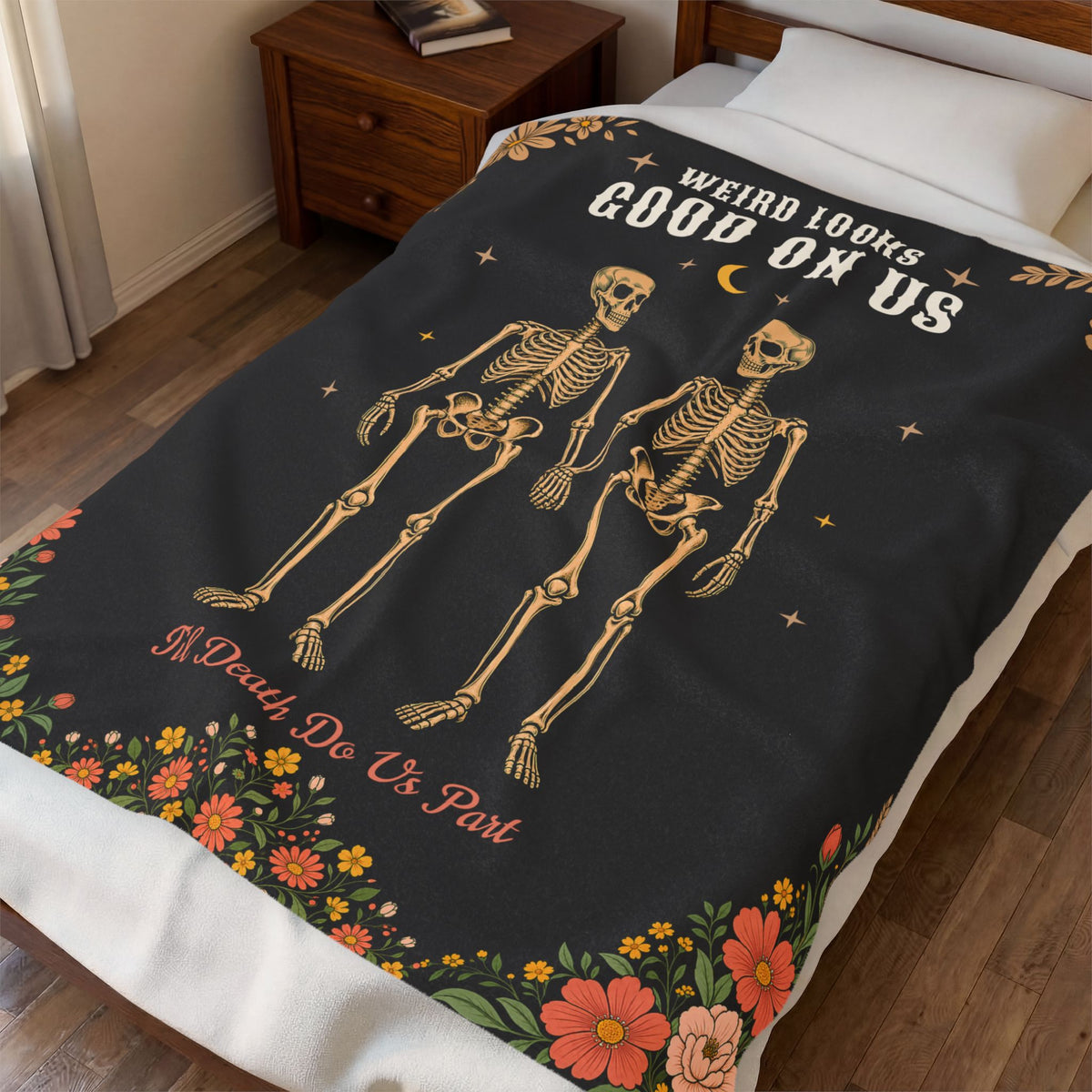 Seen from above, a Black velveteen plush blanket shows skeleton lovers holding hands on a made bed beside a wooden nightstand.