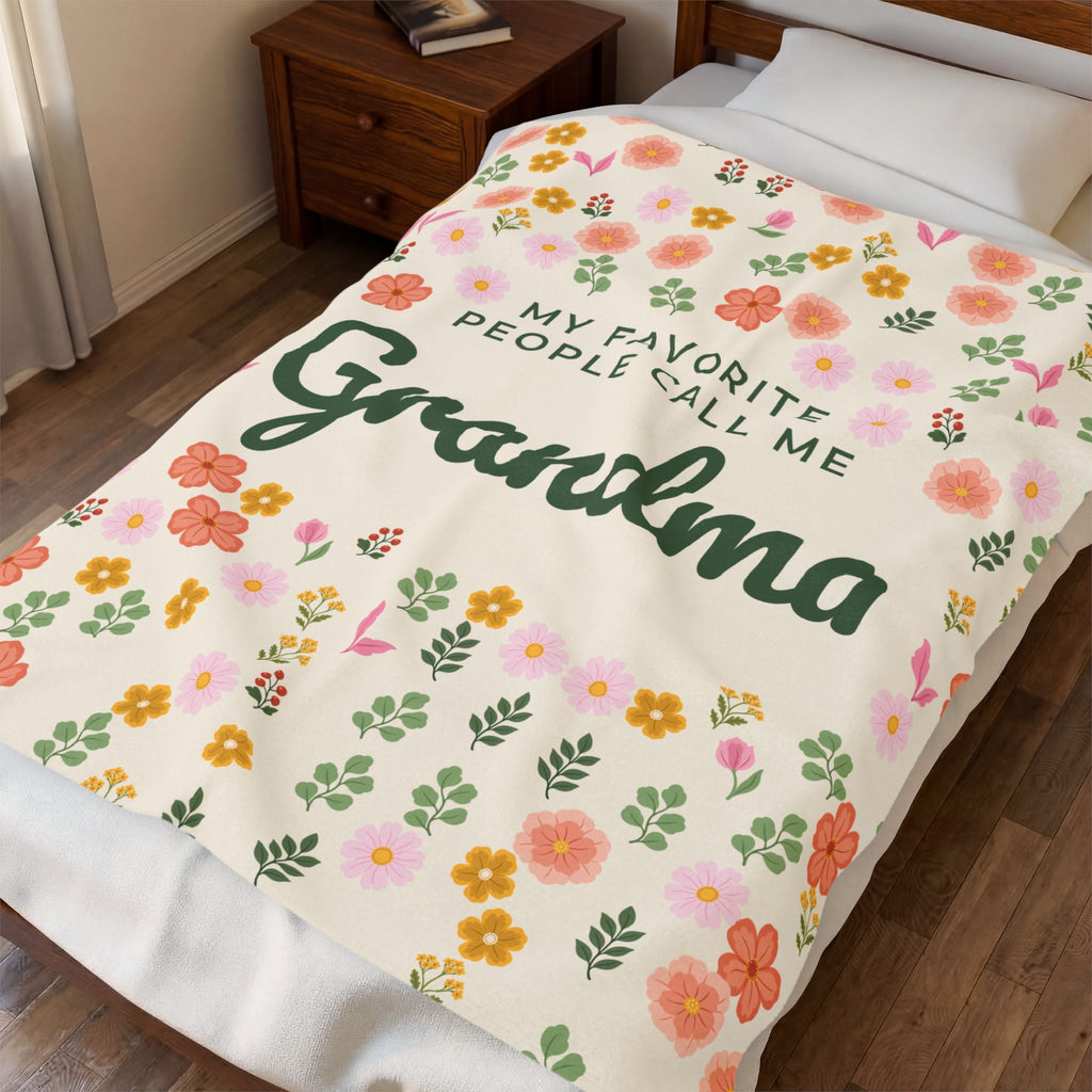 Seen from the foot of a made bed, a Cream floral blanket in velveteen plush shows forest green "Grandma" script beside a wooden nightstand.