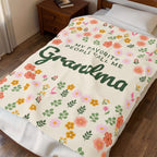 Seen from the foot of a made bed, a Cream floral blanket in velveteen plush shows forest green "Grandma" script beside a wooden nightstand.
