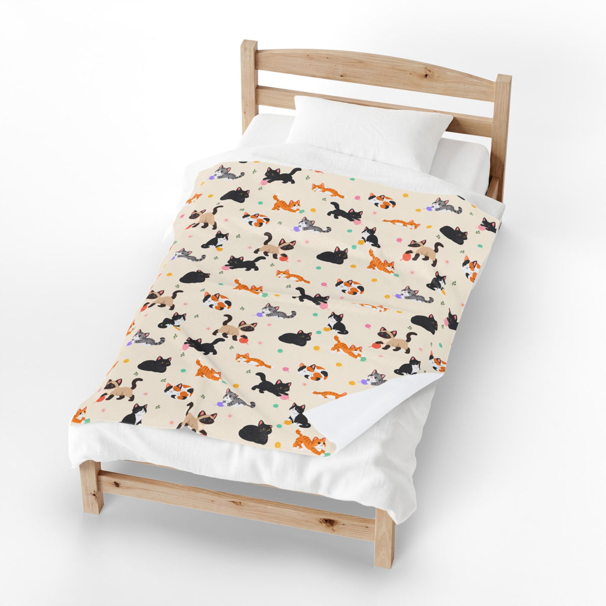 One corner of a Cream velveteen plush blanket turns back on a wooden bed, showing its plain white back beside the cartoon cats.