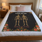 In a bright bedroom, a 50 x 60 inch Black velveteen plush blanket with tan skeletons and wildflowers lies on a white duvet.