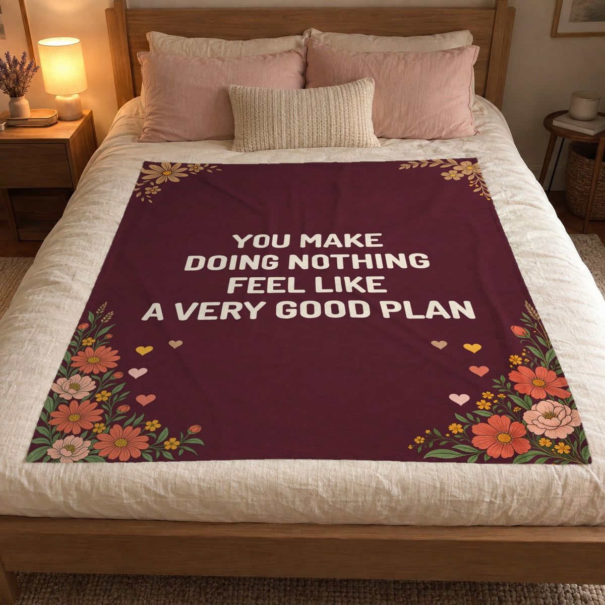 A 50 x 60 inch Wine romantic blanket in velveteen plush shows cream words on a bed with a cream duvet, blush pillows and a lit lamp.