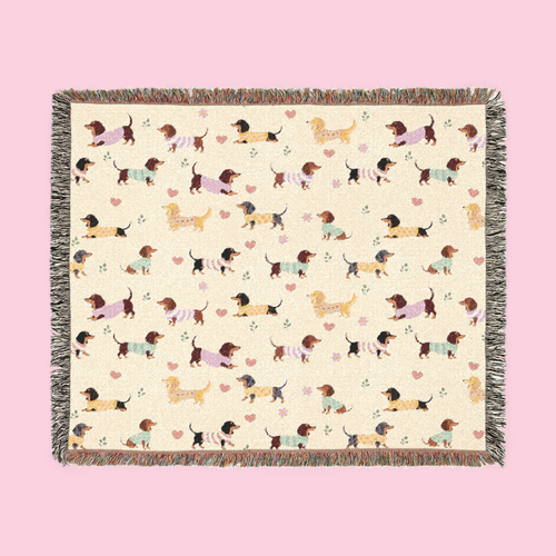 Colorful fringe borders a Cream woven cotton blanket laid flat, showing dachshunds in knit sweaters.