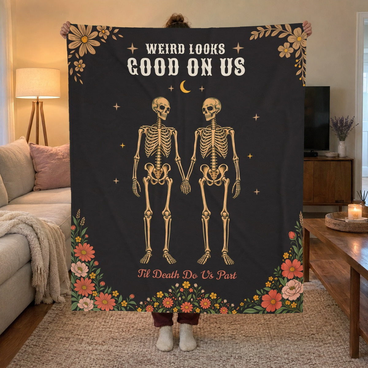 A woman holds up a 50 x 60 inch Black velveteen plush blanket with a skeleton couple in a media room with warm lamp light.