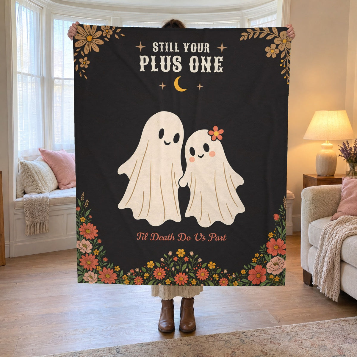 By a family room bay window, a woman holds up a 50 x 60 inch Black velveteen plush ghost blanket showing two smiling cream ghosts.