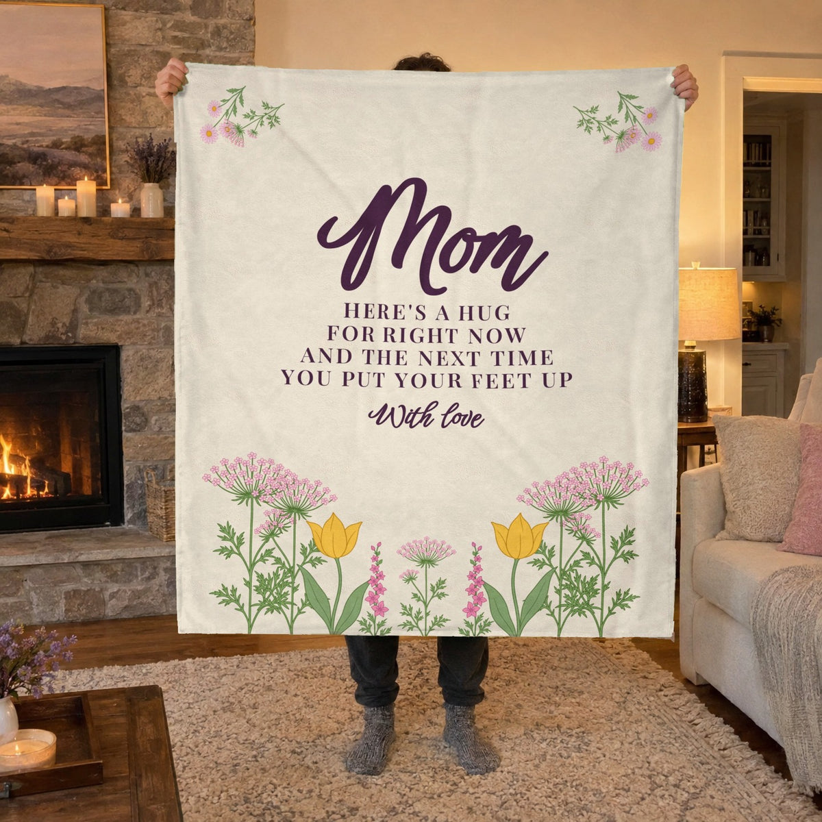 Beside a lit stone fireplace, a man holds up a 50 x 60 inch Cream velveteen plush blanket with "Mom" above a wildflower meadow.