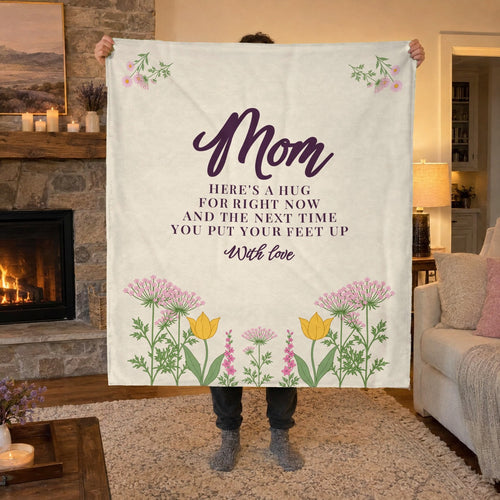 Beside a lit stone fireplace, a man holds up a 50 x 60 inch Cream velveteen plush blanket with "Mom" above a wildflower meadow.