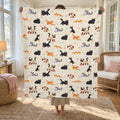 In a bright sunroom, a woman holds up a 50 x 60 inch Cream velveteen plush blanket with cartoon cats, her bare feet showing below.