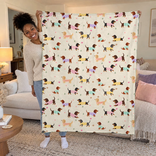 A woman holds up a 50 x 60 inch cream velveteen plush blanket with dachshunds in knit sweaters in a bright living room.