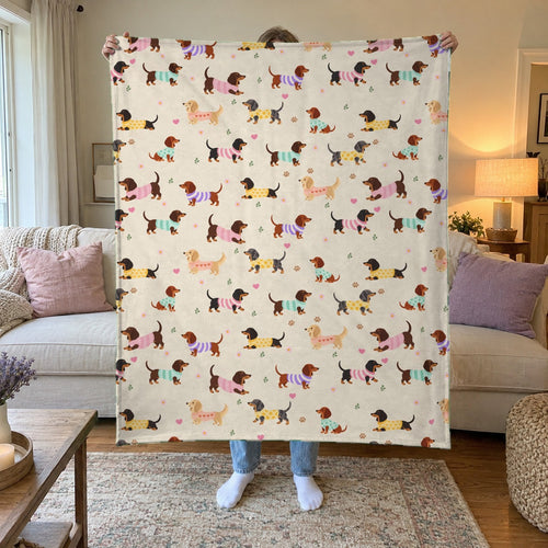 A woman holds up a 50 x 60 inch Cream velveteen plush blanket with dachshunds in knit sweaters in a living room with a cream sofa.