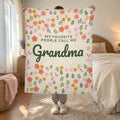 In a white bedroom with a rattan headboard, a woman holds up a 50 x 60 inch Cream velveteen plush blanket reading "MY FAVORITE PEOPLE CALL ME Grandma".