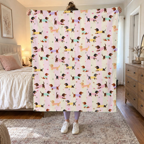 In a bedroom, a woman holds up a 50 x 60 inch Pink stripes velveteen plush blanket with dachshunds in knit sweaters beside a made bed.