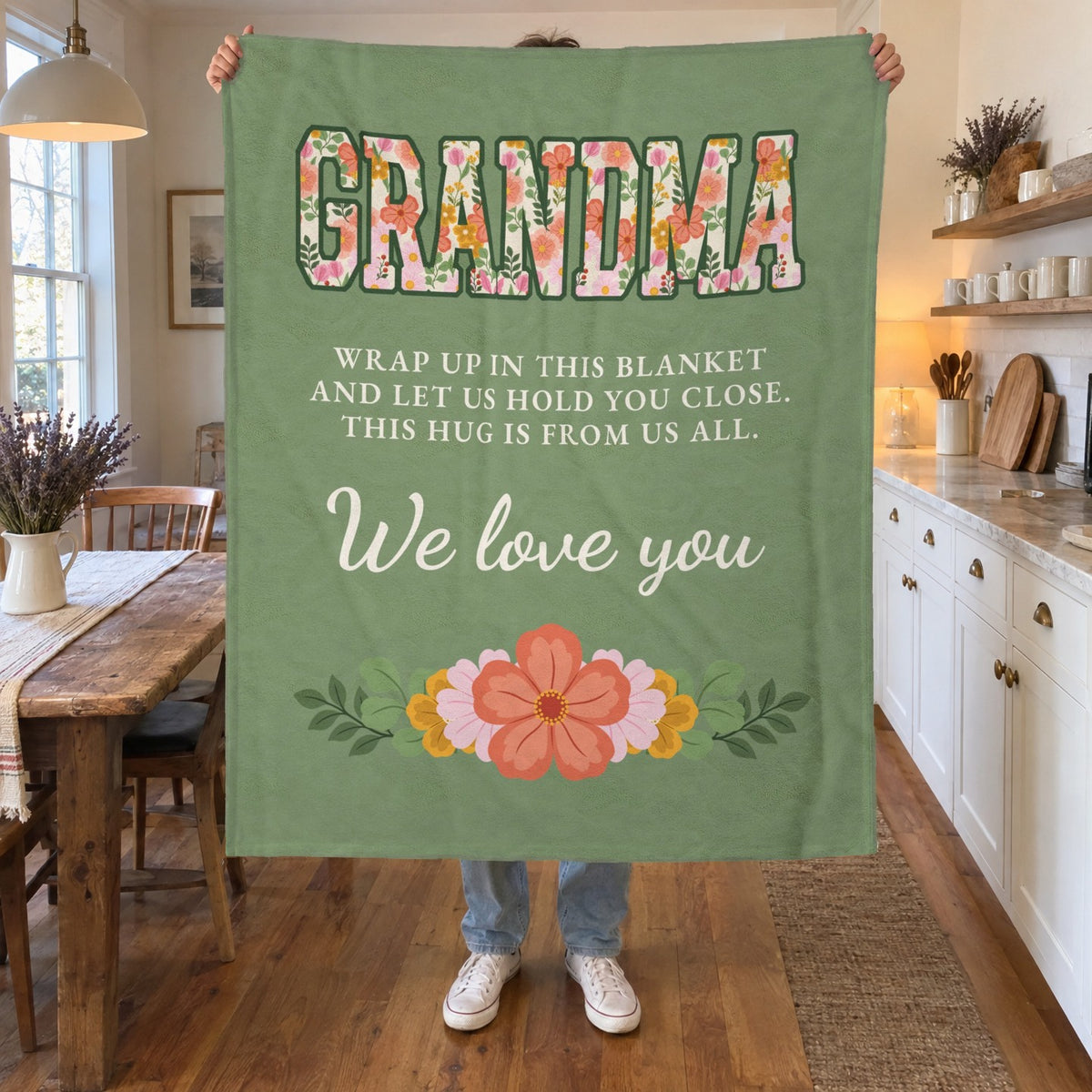 In a farmhouse kitchen with white shaker cabinets, a man holds up a 50 x 60 inch Sage velveteen plush grandma blanket with floral "GRANDMA" letters.