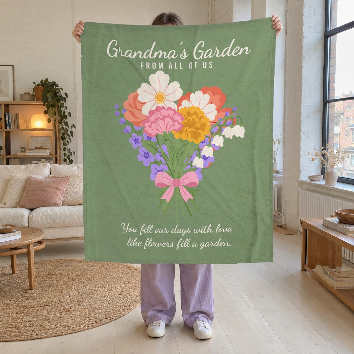 In a bright loft living room, a woman holds up a 50 x 60 inch Sage Grandma's Garden blanket in velveteen plush against white brick walls.