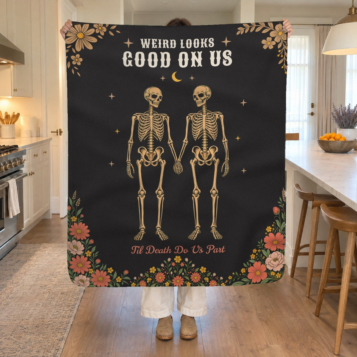 In a bright kitchen, a woman holds up a 50 x 60 inch Black sherpa fleece skeleton blanket beside a white island and wooden stools.