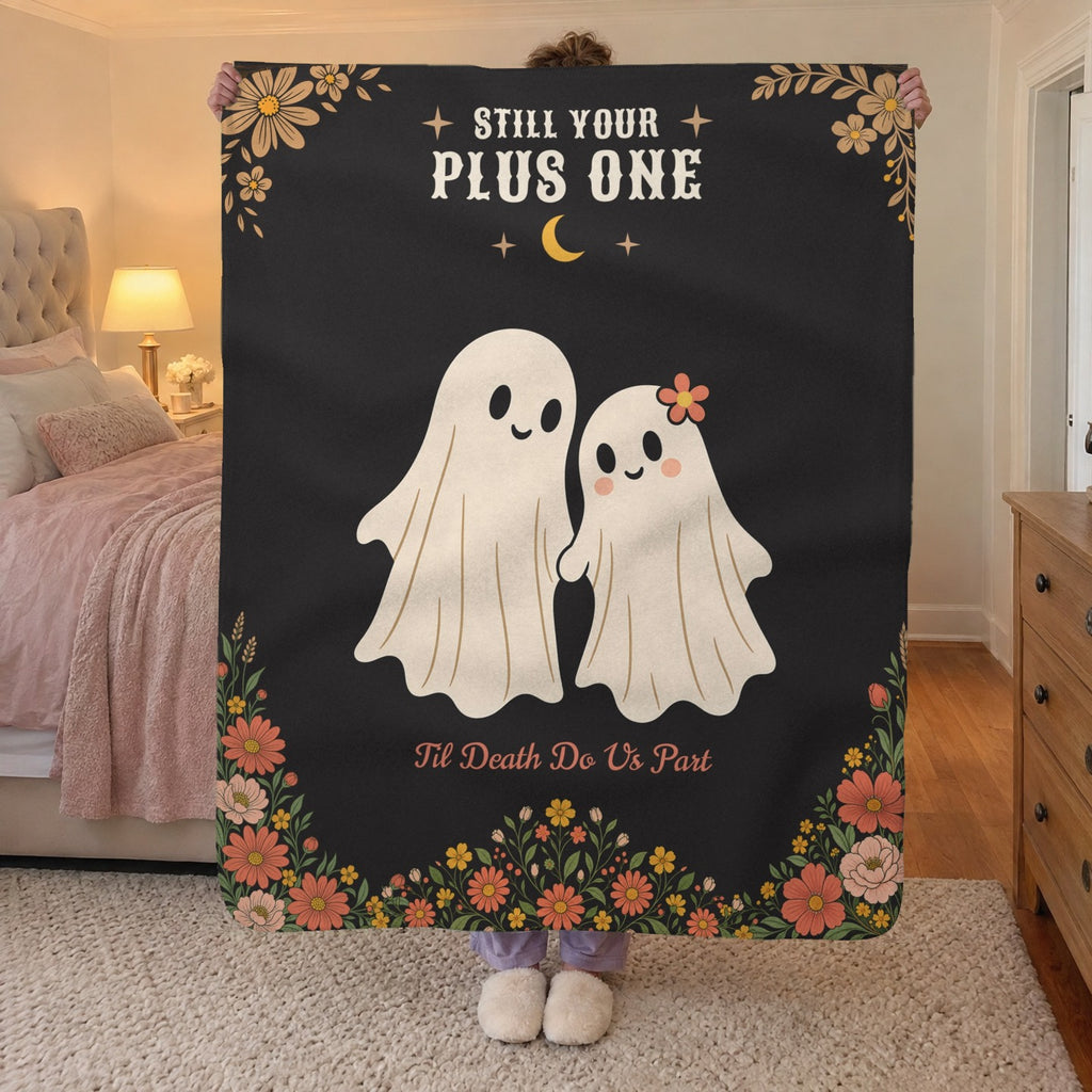 A woman holds up a 50 x 60 inch Black sherpa fleece blanket with two cream ghosts in a bedroom with blush bedding and a brass lamp.