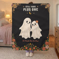 A woman holds up a 50 x 60 inch Black sherpa fleece blanket with two cream ghosts in a bedroom with blush bedding and a brass lamp.