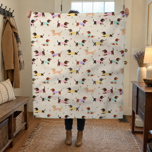 A woman holds up a 50 x 60 inch Cream sherpa fleece blanket with dachshunds in knit sweaters in a hallway with a white door and bench.