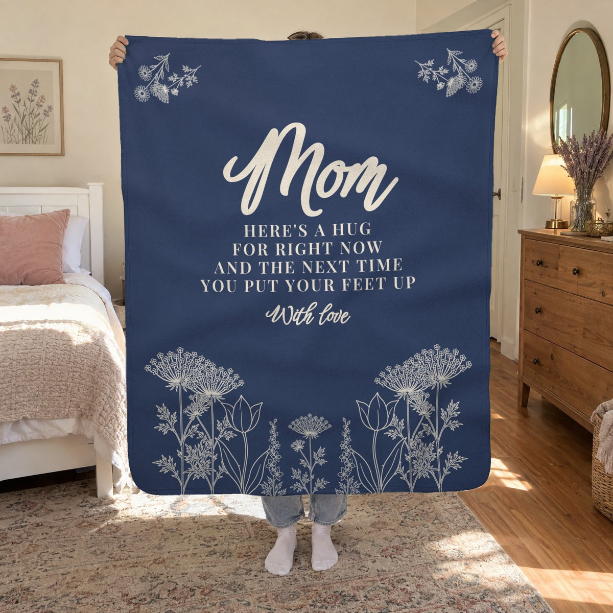 A woman holds up a 50 x 60 inch Navy sherpa fleece blanket with cream flowers in a sunny bedroom beside a white wooden bed.