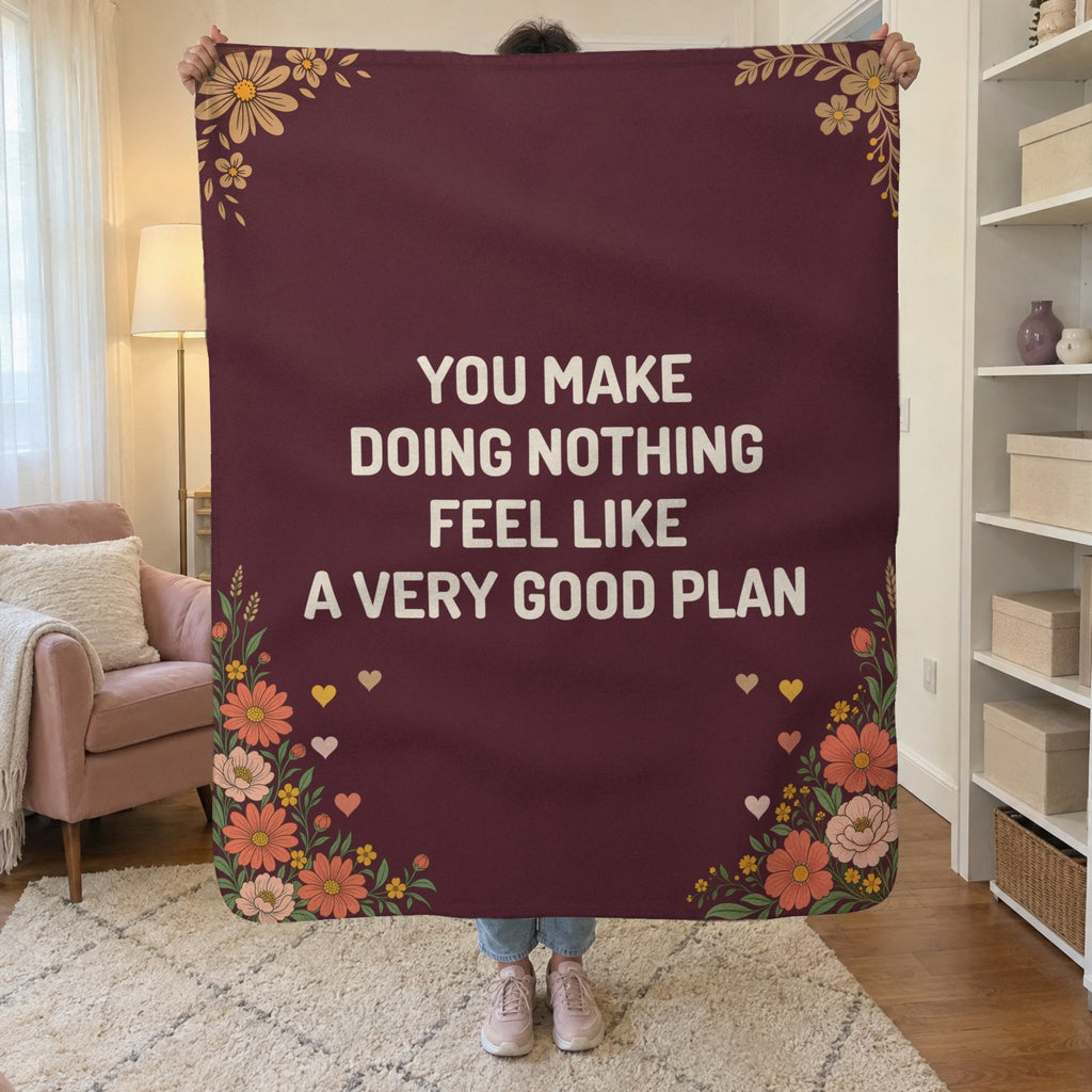 In a small living room, a woman holds up a 50 x 60 inch Wine sherpa fleece blanket with cream words beside a blush armchair.
