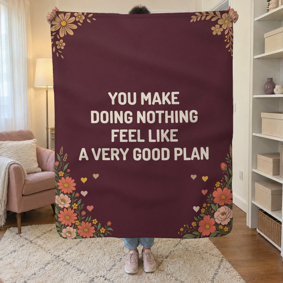 In a small living room, a woman holds up a 50 x 60 inch Wine sherpa fleece blanket with cream words beside a blush armchair.