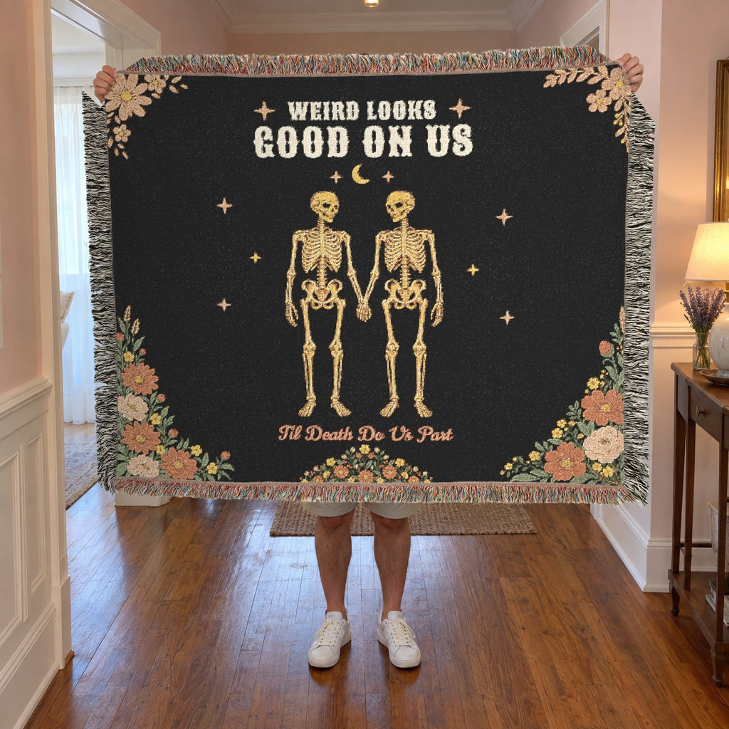 A man holds up a fringed 60 x 50 inch Black woven cotton blanket with two skeletons in a hallway with soft painted walls.