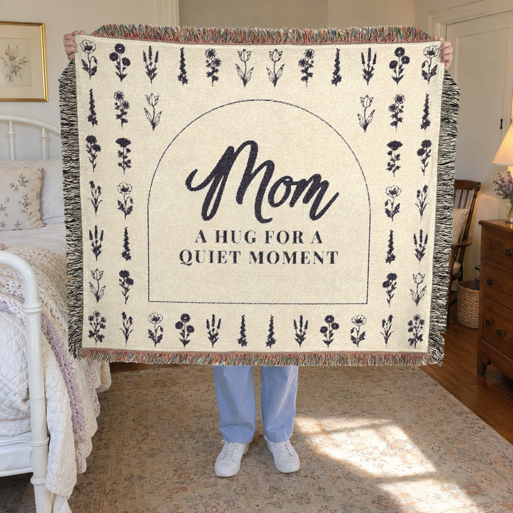 A woman holds up a fringed 60 x 50 inch Cream woven cotton blanket with a dark arch and flower rows in a sunlit guest room beside an iron bed.
