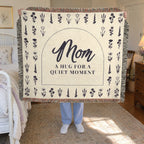 A woman holds up a fringed 60 x 50 inch Cream woven cotton blanket with a dark arch and flower rows in a sunlit guest room beside an iron bed.