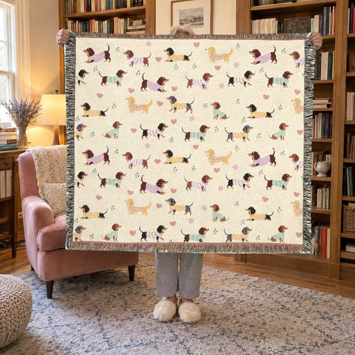 A woman holds up a fringed 60 x 50 inch Cream woven cotton dachshund blanket in a reading nook with wooden bookshelves and an armchair.