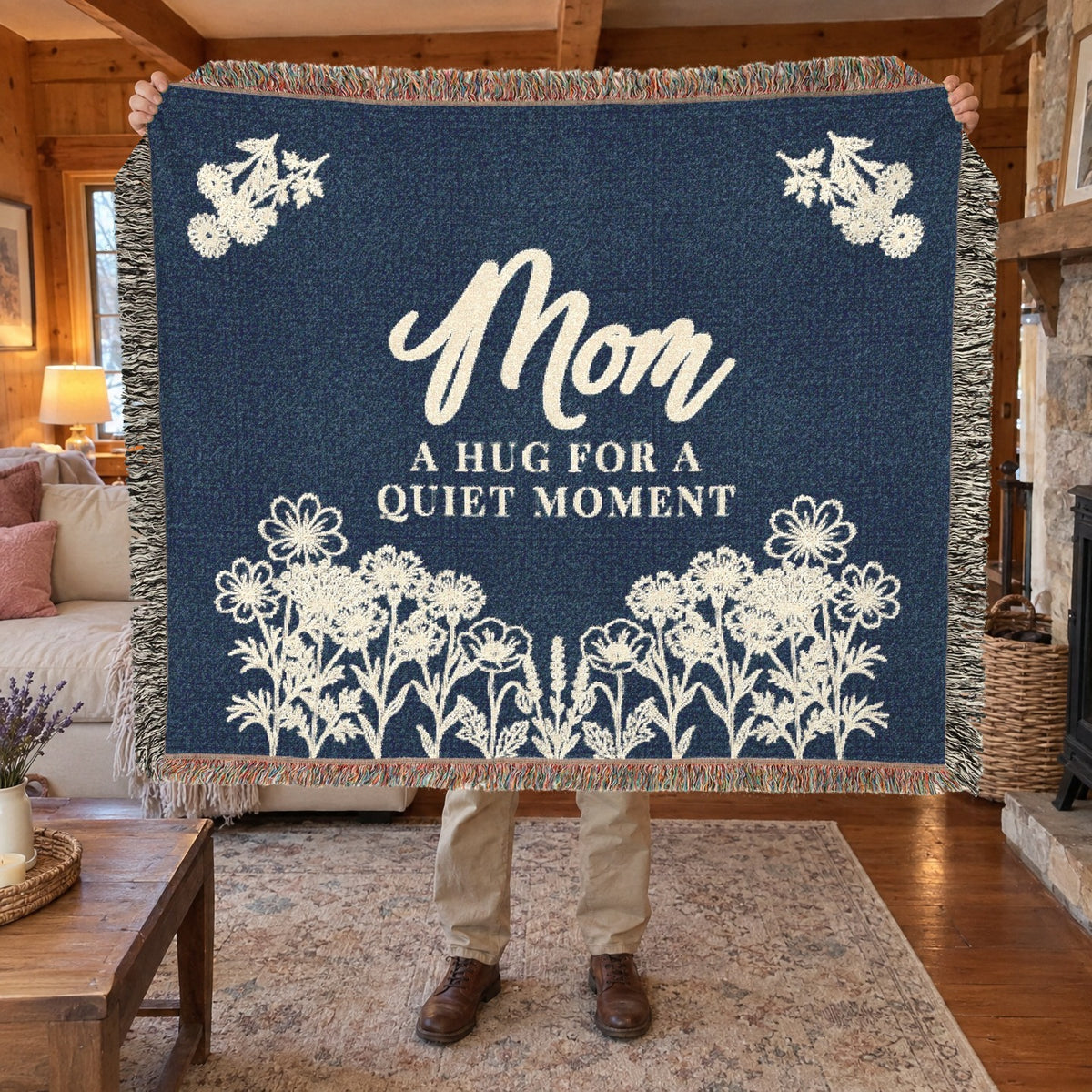 A man holds up a fringed 60 x 50 inch Navy woven cotton blanket with cream flowers in a living room with wood walls and a stone fireplace.