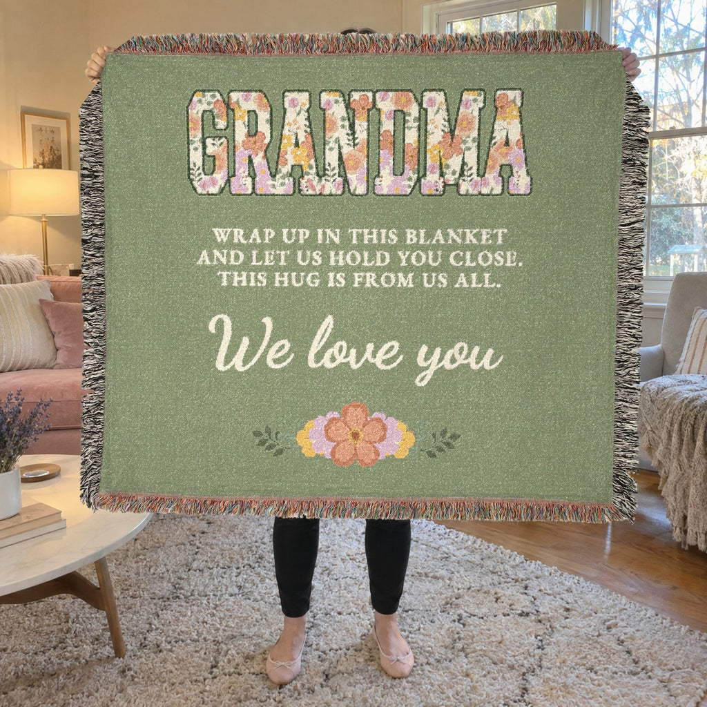 Beside a velvet living room sofa, a woman holds up a fringed 60 x 50 inch Sage woven cotton grandma throw blanket with floral "GRANDMA" letters.