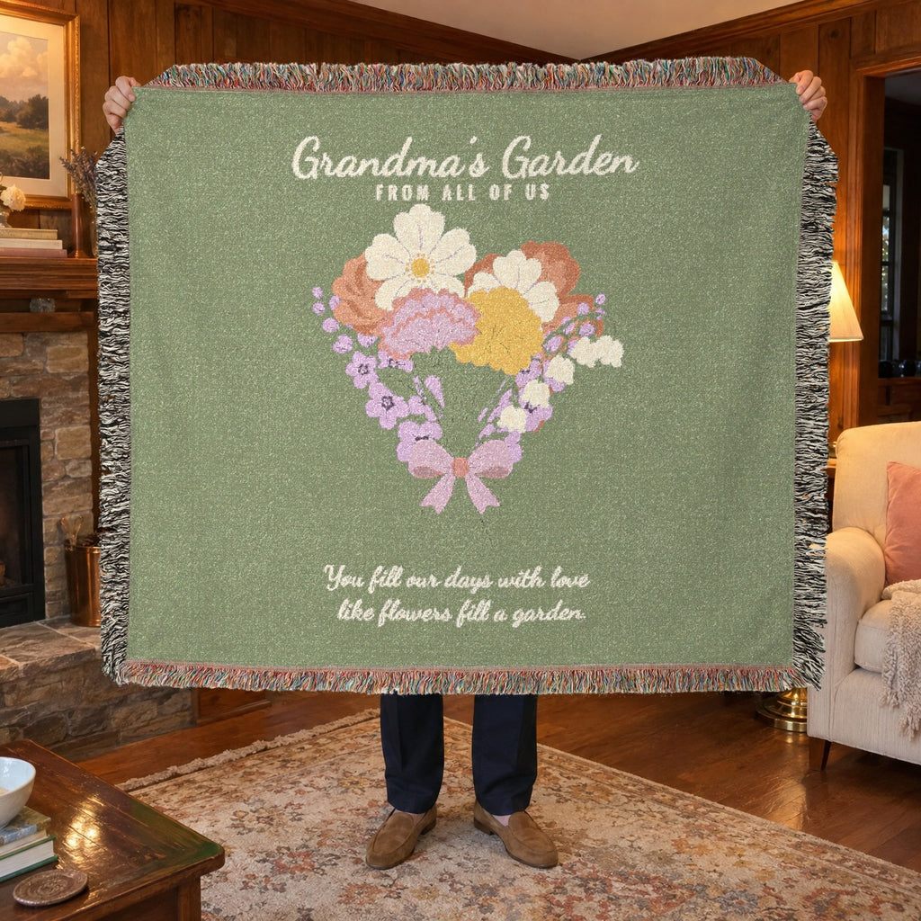 In a den with wood paneling, a man holds up a fringed 60 x 50 inch Sage woven cotton flower blanket reading "Grandma's Garden" beside a lit brass lamp.