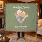 In a den with wood paneling, a man holds up a fringed 60 x 50 inch Sage woven cotton flower blanket reading "Grandma's Garden" beside a lit brass lamp.