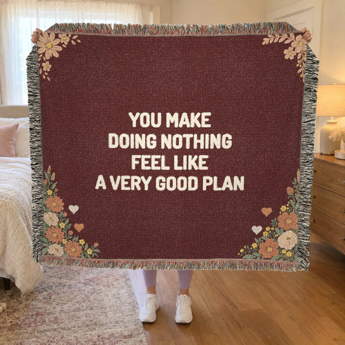 A woman holds up a fringed 60 x 50 inch Wine woven cotton anniversary blanket with cream words in a bedroom with cream bedding.