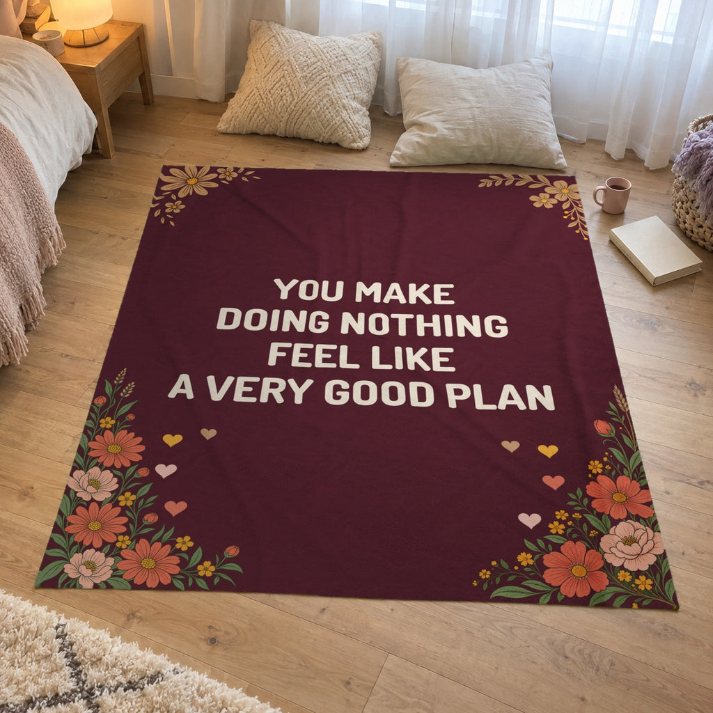 Beside a bedroom window, a 50 x 60 inch Wine velveteen plush love letter blanket with cream words lies on a wooden floor by a book and mug.